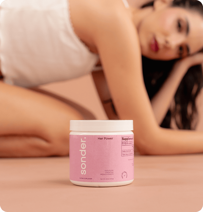 A pink container of Sonder “Her Power” 100% pure creatine monohydrate is placed in the foreground, with a woman in a white outfit kneeling and leaning to the side in the softly blurred background.