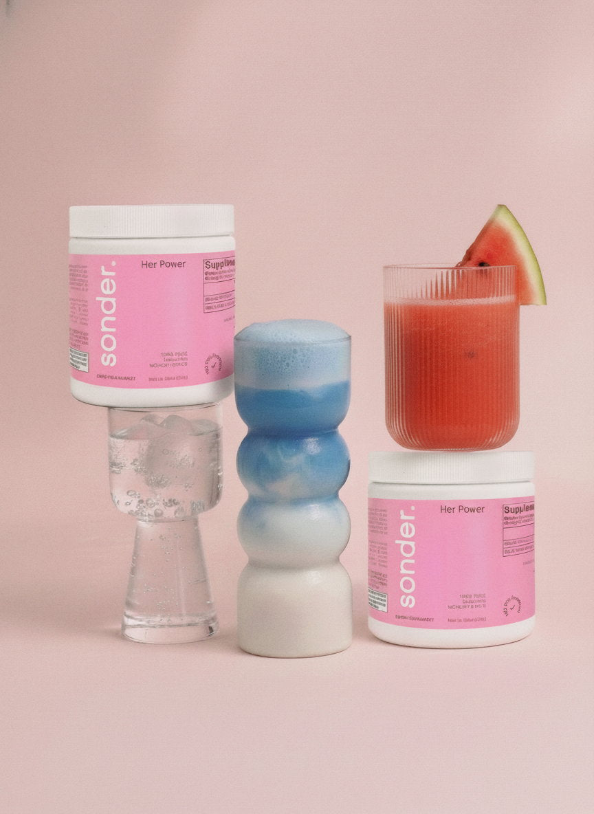 Two pink Sonder containers of 100% pure creatine monohydrate labeled “Her Power” are displayed alongside a clear fizzy drink, a blue layered beverage, and a red juice with a watermelon slice, all arranged against a soft pink background.